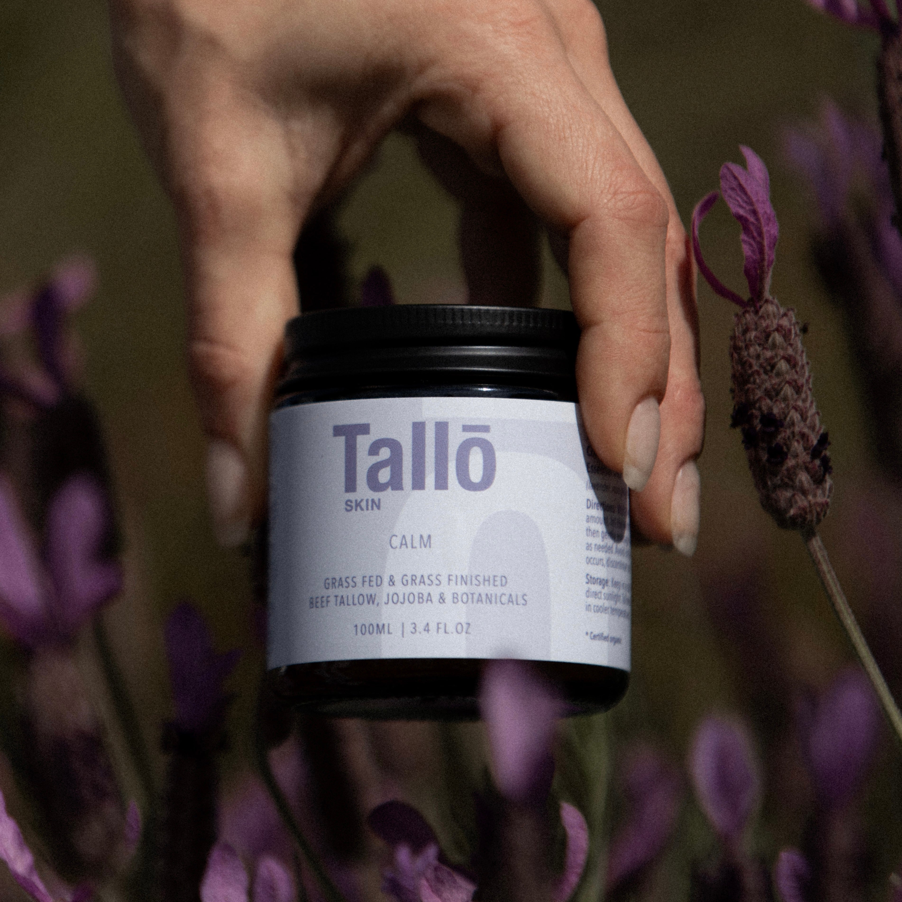 Hand holding a jar of Tallo skincare product with lavender flowers in the background